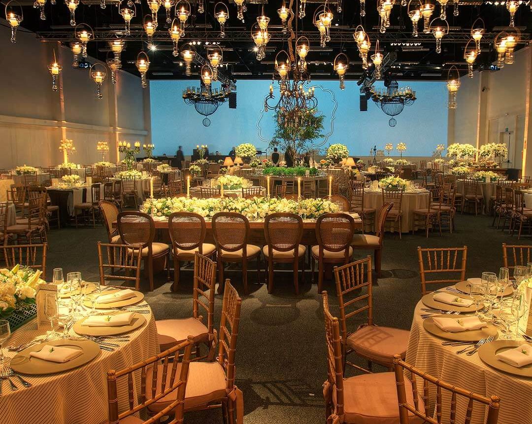 Salão de eventos, com diversas mesas, cadeiras, lustres, flores, telão ao fundo e outros itens no ambiente